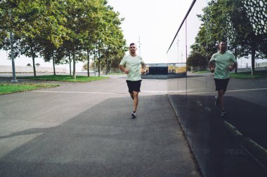 Motivasyonlu erkek koşucu sabah vakti aktif giyinip koşuyor sportif yaşam tarzından zevk alıyor, eşofmanlı yakışıklı adam şehirde fiziksel koşu için enerjik bir kardiyo eğitimi alıyor.