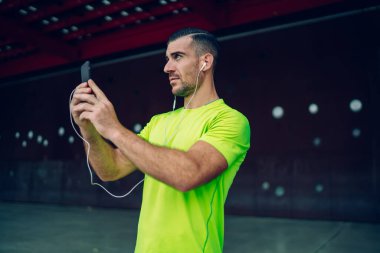 Elektronik kulaklıklı genç beyaz sporcu motivasyonlu ses kitabı dinliyor ve video vlog çekmek için mobil kamera kullanıyor. Kulaklıklı kaslı erkek koşucu şehir merkezlerini fotoğraflıyor.