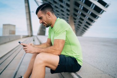 Kafkasyalı genç sporcunun cep telefonu ağları sırasında motivasyon yayını okumasının yan görüntüsü. Kaslı erkek atlet cep telefonu teknolojisinden 4G kablosuz bağlantı ile e-posta alıyor.