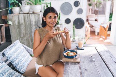 Kısa siyah saçlı hoş bir kadın güneşli terasta masada oturuyor ve öğle yemeği sırasında kameraya bakarken akıllı telefon kullanıyor.