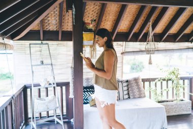 Bali tatil beldesinde terasta dikilen ve tatilde akıllı telefonları karıştıran gündelik giysiler içindeki neşeli etnik kadın.