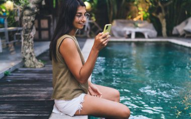 Yaz tatili boyunca havuz başında otururken günlük kıyafetlerle gezen mutlu etnik kadın manzarası