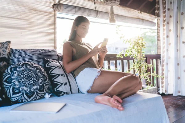 Gün ışığında aydınlık odada akıllı telefon kullanırken rahat koltukta dinlenen çıplak ayaklı kadının yan görüntüsü.