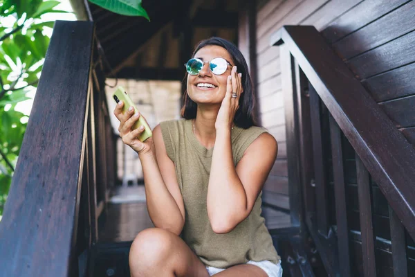 Sıradan giyinmiş, güneş gözlüklü, ahşap merdivenlerde dinlenen ve Bali 'de dinlenirken akıllı telefondan sohbet eden mutlu bir kadın.