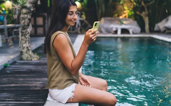 Yaz tatili boyunca havuz başında otururken günlük kıyafetlerle gezen mutlu etnik kadın manzarası