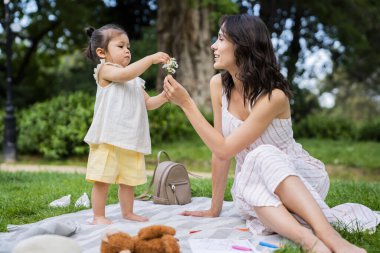 Siyah saçlı, neşeli, çizgili günlük elbiseli bir kadın parkta battaniyede oturuyor ve küçük çiçekleri sevimli bebeğe veriyor.