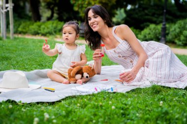 Koyu renk saçlı, neşeli bir kadın. Parkta battaniyede dinleniyor. Balon üfleyicisine merakla bakan küçük bir kızla birlikte.