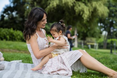 Dikkatli bir annenin gülümsemesi ve şirin bir çocuğu dizlerinin üstünde battaniyeye yatırması parkta piknik yaparken. 