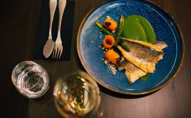 Yeşil soslu seramik tabakta servis edilen kuşkonmaz ile birlikte deniz levreği filetosundan yapılmış lezzetli yemeğin üst görüntüsü ve gümüş çatal bıçak ve içeceklerin yanındaki masaya yerleştirilmiş.