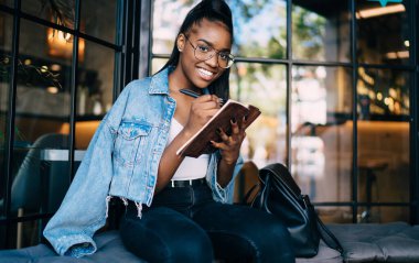 Gözlüklü Afro-Amerikan 'ın yarım boy portresi kafenin içinde oturmuş proje için notlar alıyor şık giyinmiş güzel siyahi kadın elinde çizimlerle kameraya bakıyor.