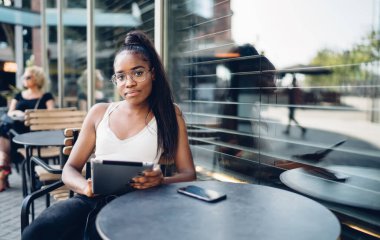 Gündelik giyinmiş yeniyetme kız portresinin yarısı boş zamanlarında dijital tablet kullanan Afro-Amerikan kadın blogcu touchpad 'i tutuyor ve kafe terasında kameraya bakıyor.