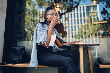 Genç modaya uygun giyinmiş güzel Afro-Amerikan hippi kız açık havada kahve içiyor, çekici, esmer tenli öğrenci kafeterya terasında elinde içecek bardağıyla mola veriyor.