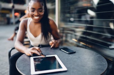 Genç gülümseyen, at kuyruklu, gözlüklü, tabletle mesajlaşan Afro-Amerikan kadın ara verdiğimizde elinde akıllı telefonuyla sokak kafesinde oturuyor.