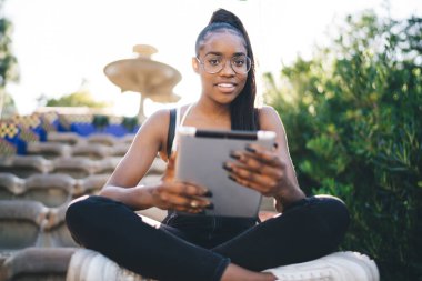Kitap okumak için modern dijital tablet taşıyan günlük giysili neşeli, koyu tenli bir kadının yarım boy portresi. Güzel Afrikalı Amerikalı hippi kız parkta gezmek için touchpad kullanıyor.