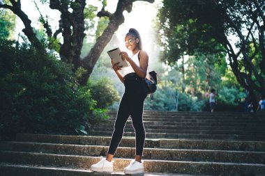 Parktaki merdivenlerde dururken tablet kullanarak günlük giysiler ve gözlük takan Afro-Amerikan genç bayanlardan.