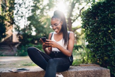 Sıradan giyinen mutlu Afrikalı Amerikalı kadın sosyal ağlarda cep telefonuyla mesajlaşıyor, gülümseyen güzel bir kadın parkta eğleniyor ve blog ve web sayfasına göz atmak için 4G bağlantısı kullanıyor.