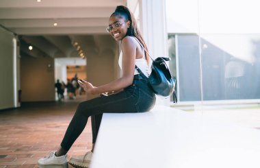 Modern koridorda tablet ve sırt çantasıyla oturan sıradan giysili ve gözlüklü mutlu Afrikalı genç bir Amerikalı kadın.