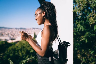 Mutlu, uzun saçlı, yaz kıyafetleri ve gözlüklü, güneşli bir günde parkta akıllı telefon kullanan siyah kadının yan görüntüsü.