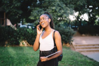 Gözlüklü ve tabletli Afro-Amerikan bir kadının parkta durup komik telefon konuşmaları yaparken gülüp başka tarafa bakması.