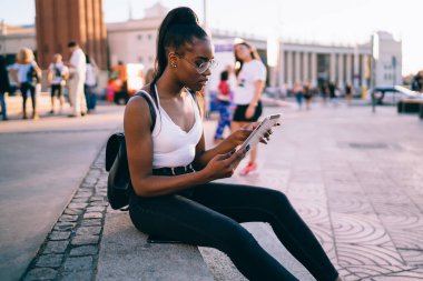 Gözlüklü, sırt çantalı, caddede merdivenlerde oturan ve hafta sonu dinlenirken tablet okuyan Afrikalı genç bir kadının yan görüntüsü.