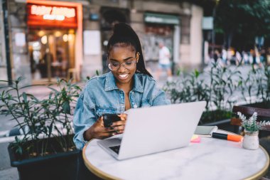 Keyifli genç Afrikalı Amerikalı kadın ara verildiğinde açık kafede dizüstü bilgisayarla dinlenirken gözlüklü cep telefonu takıyor.
