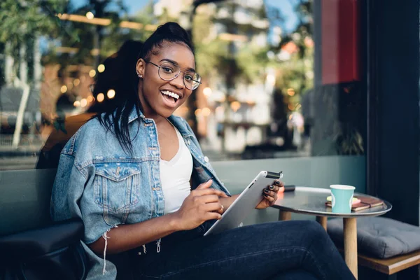 穿着时髦眼镜的黑人女性穿着正装 一边在平板电脑上冲浪 一边在咖啡店里喝咖啡 一边用张开的嘴看着相机 — 图库照片