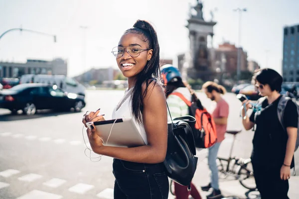 非洲裔美国妇女在靠近道路的现代城市街道上漫步时使用平板电脑和耳机的侧视图 — 图库照片