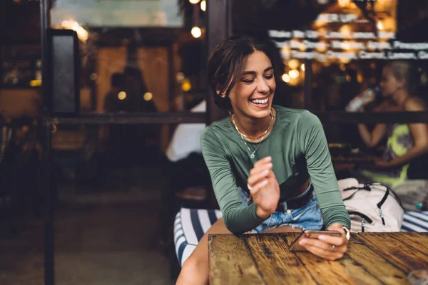 Optimistic female in casual outfit smiling and chatting on mobile phone while resting at wooden table in cozy cafe during weekend