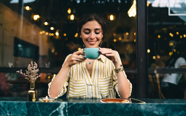 坐在舒适的街边咖啡店外的桌上 穿着休闲装的年轻女性快乐地微笑着 一边喝着热咖啡 — 图库照片