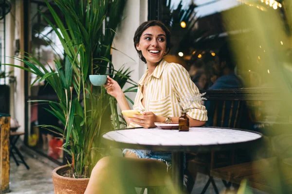 身着休闲装的年轻女性微笑着 一边喝着咖啡 一边用手机 一边坐在小圆桌边 一边在街上的咖啡店里享受着时光 — 图库照片