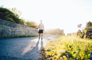 Tanımlanamayan, sıradan giyinmiş, yaz mevsiminde akşam güneşine karşı boş kaldırım taşı patikasında dikilen bir erkek vücudu.