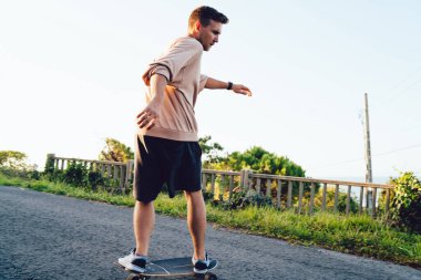 Sıradan giyinen beyaz erkek kaykaycı, kırsal alanda pratik yapıyor ve paten kayıyor, yakışıklı hippi uzun panoda antrenman yapıyor, asfalt yolda yaz tatillerinin keyfini çıkarıyor.