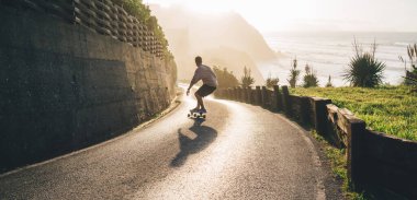 Kırsal kesimde asfalt yolda kayak yapan ve pratik yapan profesyonel erkek kaykaycı, günbatımında, serbest giyinen genç hippi çocuk yaz tatillerinde serbest stil paten yapıyor.