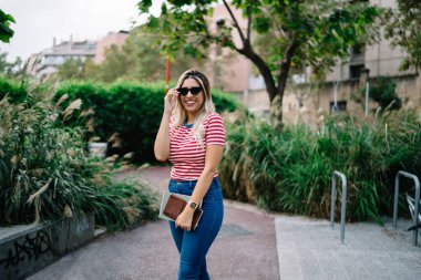 Moda giyinmiş, güneş gözlüğü takmış, sokakta poz veren neşeli beyaz kadın portresi. Güzel hippi kız, şehir parkının arka planında poz veriyor.