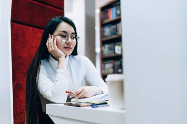 Çekici düşünceli Asyalı kadın kitap okuyor ve masaya oturmuş modern kitapçıda elini uzatıyor.