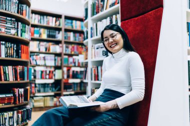 Gözlüklü neşeli Asyalı kadının modern sandalyede oturup kitap okuyarak gülüşü ve kütüphanede kameraya bakışı.