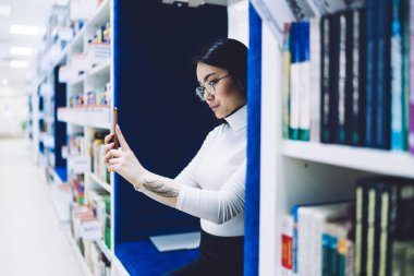Kolunda dövmesi olan, cep telefonuyla fotoğraf çeken, kütüphanedeki kitaplıklar arasında oturan Asyalı genç bir kadının yan görüntüsü