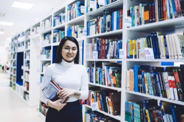 Koyu renk saçlı, neşeli Asyalı, gözlüklü, sıradan giyinmiş, elinde kitap tutan ve modern tasarım kütüphanesinde duran bir kadın. 