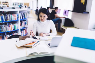 Büyük kütüphanede dizüstü bilgisayarda çalışırken kitaplarda bilgi arayan gülümseyen Asyalı öğrenci kameraya bakıyor.