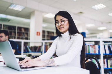 Genç, kendinden emin, koyu renk saçlı, gözlüklü ve elinde dövmesi olan bir kadın kütüphanede dizüstü bilgisayarında yazarken kameraya bakıyor.