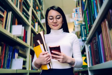 Beyaz balıkçı yakalı siyah saçlı genç Asyalı kadın ve kitaplarla dolu gözlüklü akıllı telefon kütüphanede rafların arasında duruyor.