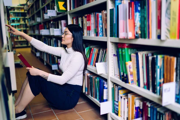 亚洲女学生坐在地板上 手里拿着书 靠着书架 在大学图书馆里找文学的侧视图 — 图库照片