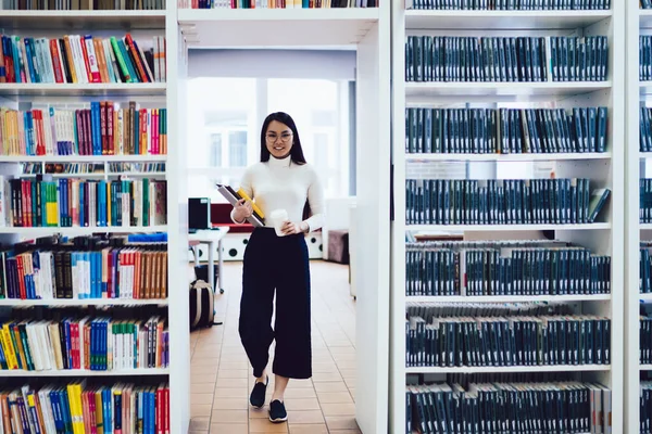 在轻图书馆的书架上 穿着黑色长裤 白色高领毛衣 头戴笔记本 头戴白色一次性咖啡杯的聪明的亚洲女人站在过道里 — 图库照片