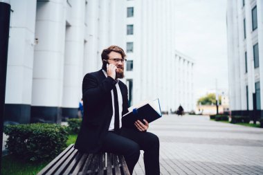 Sakallı, kendine güvenen, resmi siyah elbiseli bir işadamı boş bir caddede oturuyor, telefonda konuşurken evrak işleriyle uğraşıyor ve gözlerini kaçırıyor.