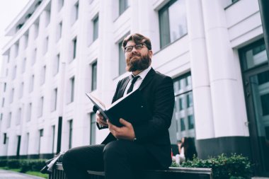 Kendine güveni tam başarılı bir işçiden resmi olarak sakallı, bankta oturan ve başka tarafa bakarken dokümanları okuyan.