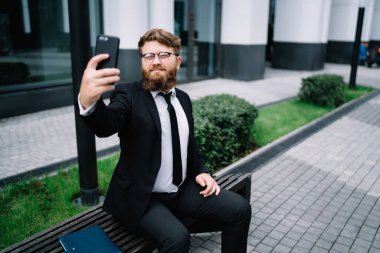 İyi giyimli, gözlüklü ve sakallı genç girişimci ofis binasının bahçesinde bankta oturuyor ve coşkuyla selfie çekiyor.