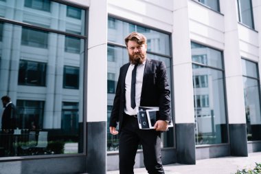 Ciddi sakallı iş adamının resmi kıyafetleri ve kravatları modern binanın yanında duruyor ve elinde belgelerle kameraya bakıyor.