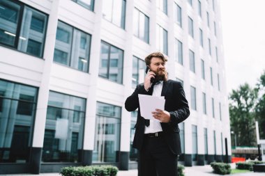 Ciddi sakallı, resmi giysiler içinde, cep telefonuyla konuşan ve modern ofis binasının yanında duran ciddi bir iş adamının alçak açısı.