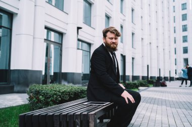 Resmî takım elbiseli dalgın sakallı adam çok katlı binanın bahçesinde dinleniyor ve yüzünü çevirmiş bakıyor.