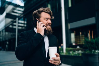 Modern binanın yakınında yürürken cep telefonuyla konuşan ve kahve içen modaya uygun resmi kıyafetli dalgın sakallı adam.